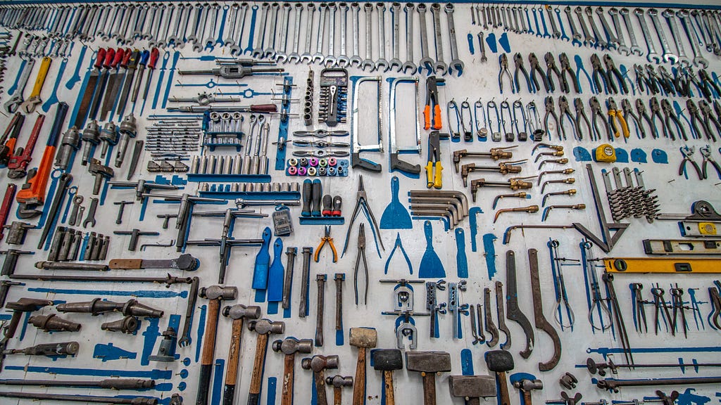 A vast array of hundreds of carefully placed hand tools, wrenches, mallets, pliers, saws, drillbits, etc. sprawls across a clean, solid, light-colored background.