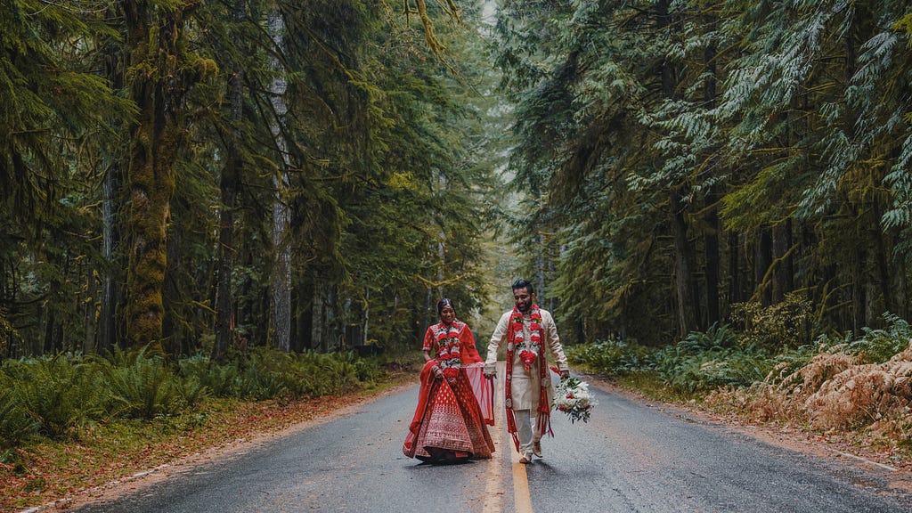 Young Indian couple in traditional sagai attire, walking down tree-flanked road in Pacific Northwest