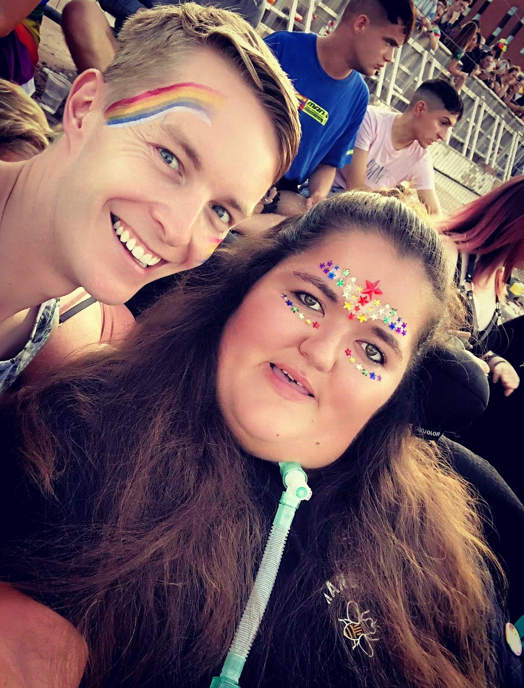 A man and woman smile at the camera. Both have rainbow paint and glitter on their faces. The woman is a wheelchair user.