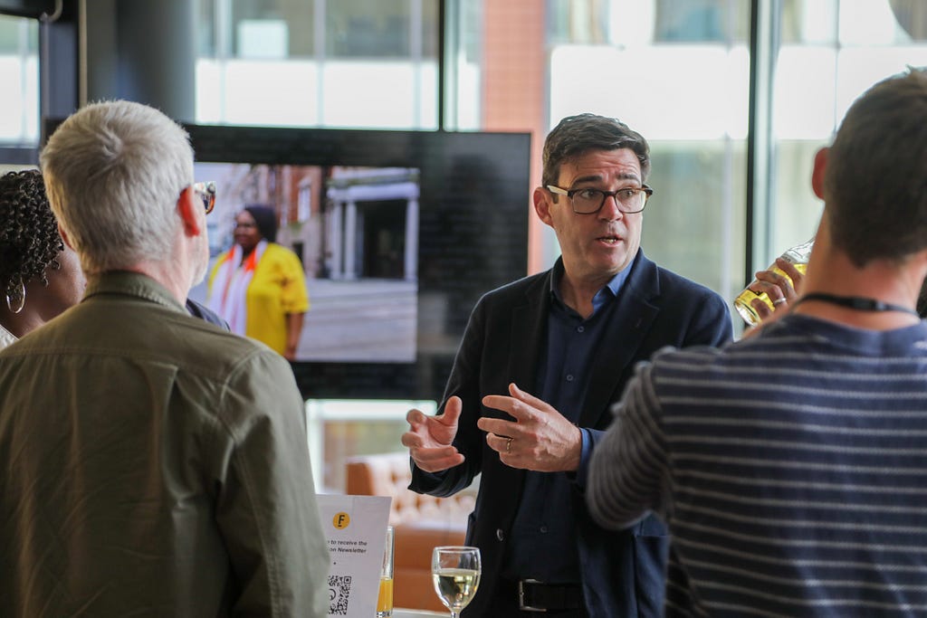 Greater Manchester Mayor, Andy Burnham is in the centre of the picture. He is talking with 4 people Federation partners who are gathered around him. They are standing in front of a TV screen which is showing an image of a woman standing in front of Federation House.