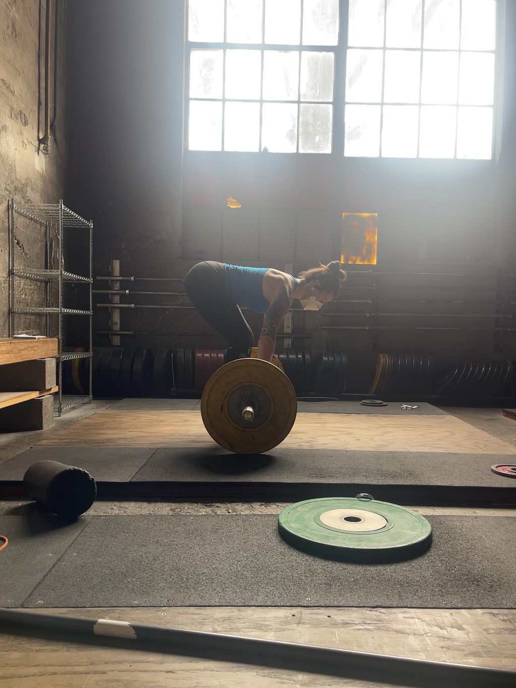The writer, Kelsey, a white woman, is seen from the side setting up at a loaded barbell on a weightlifting platform. The overhead lights are off and natural light pours in from windows at the top of the wall in the background. She’s wearing weightlifting shoes, leggings, a tank top, and a 3M Aura respirator.