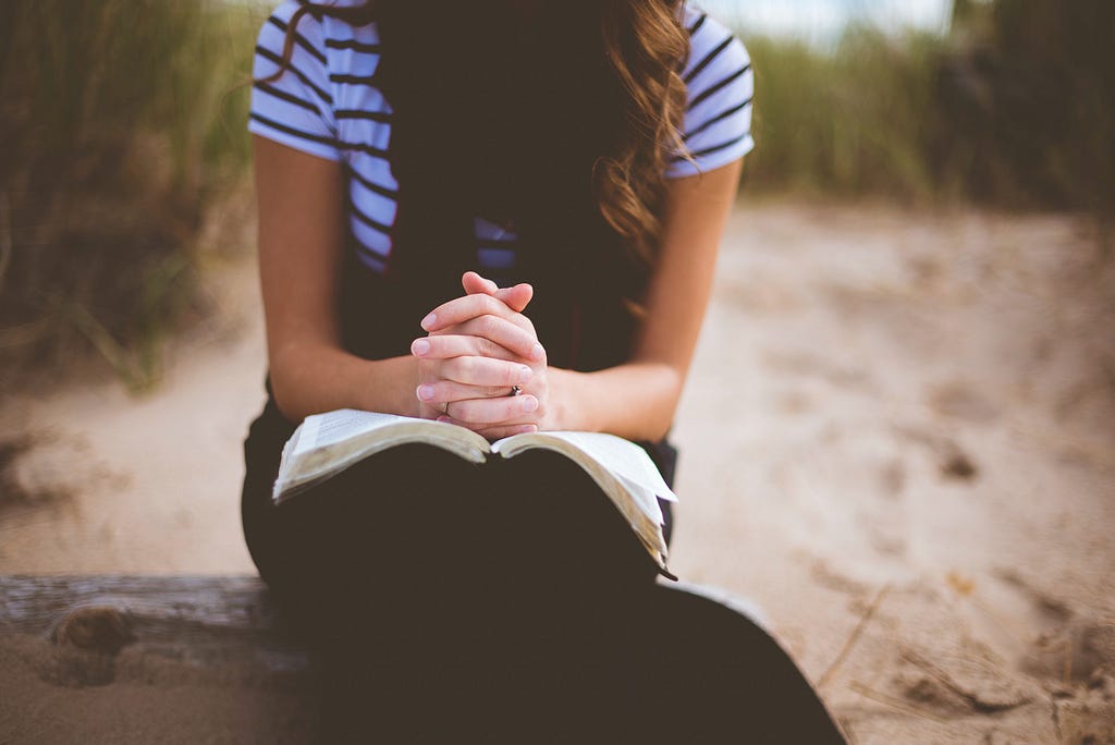 A woman is sat on a beach, head bowed down and her hands clasped as in prayer, leaning on the bible opened on her lap.