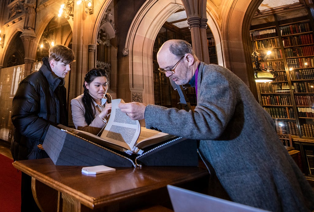 Two visitors examining a facsimile of the Gutenberg Bible, assisted by a staff member in the Historic Reading Room of the John Rylands Library