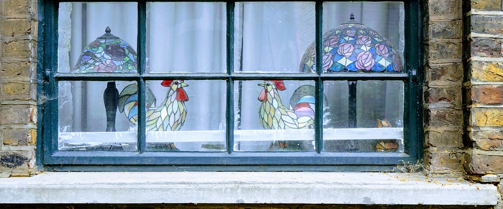 Image: two stained glass rooster sculptures facing each other, with lamps behind each one, viewed through a window pane.