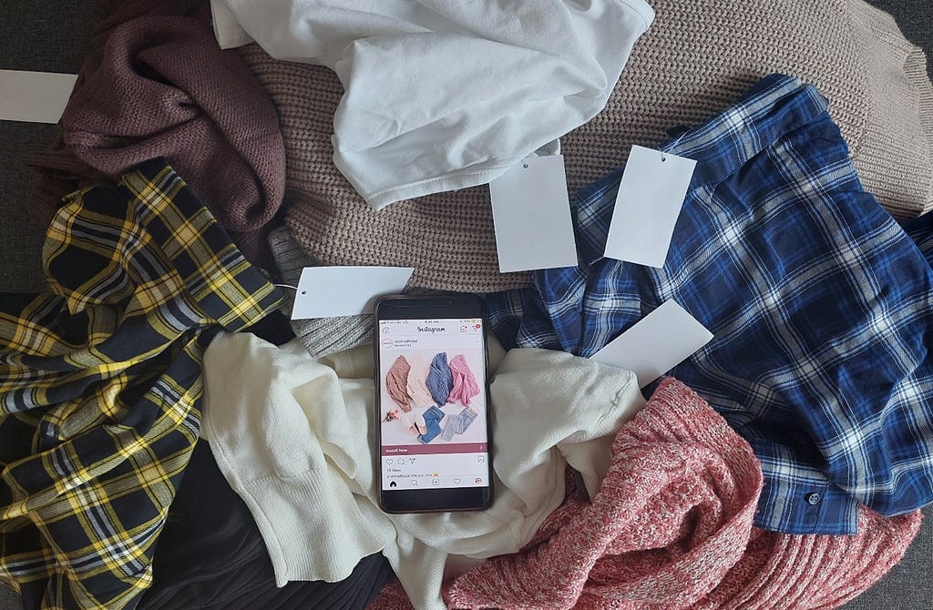 Assortment of new clothes that have tags with a smart phone in the middle showing a Shein ad as displayed on Instagram
