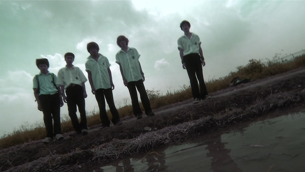 A screen capture of 5 teen boys, dressed in school uniforms that consist of white button down shirts and black pants. they are standing outside in front of a muddy pond looking down at something in the pond, on a really cloudy day. Four of teen boys stand together in a line to the left of the screen and one stands to the far right.