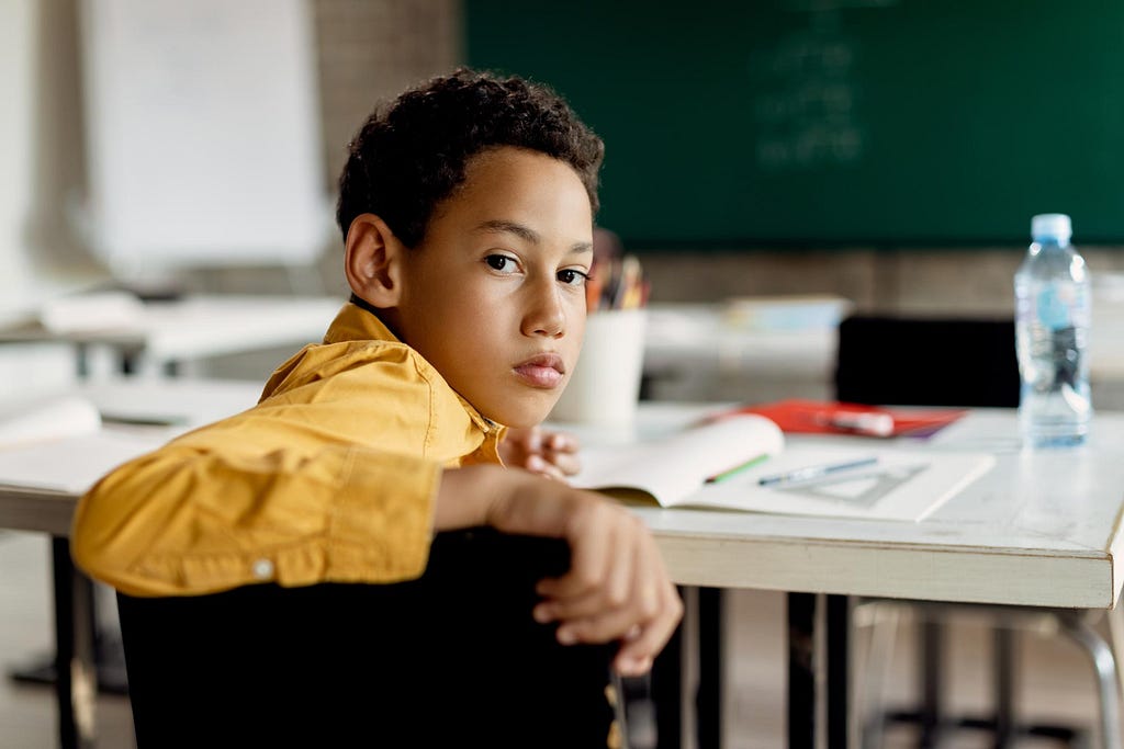 Um garoto sério está sentado em uma cadeira na sala de aula virado para trás, olhando diretamente para a câmera. Ao fundo, está uma mesa com seus materiais escolares e um quadro-negro.