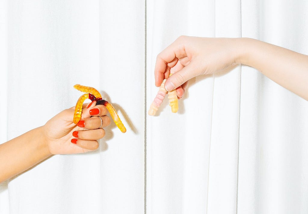 Two hands hold gummy worms over a white background.