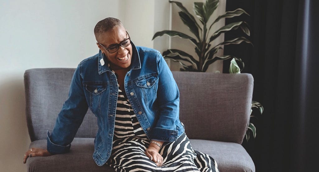 woman wearing zebra striped dress and jeans jacket, sitting on a grey couch laughing