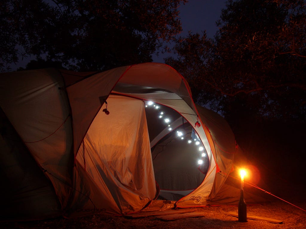 A tent at night, with fairy lights illuminating the inside, while a candle stuck in a wine bottle burns outside.