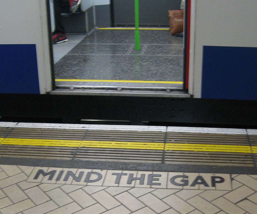 A photograph taken at a London Underground station, showing the edge of the train platform where a train’s door is open. Behind the tactile platform-edge surface, the tiles read “Mind The Gap”, referring to the gap between the platform edge and the train.