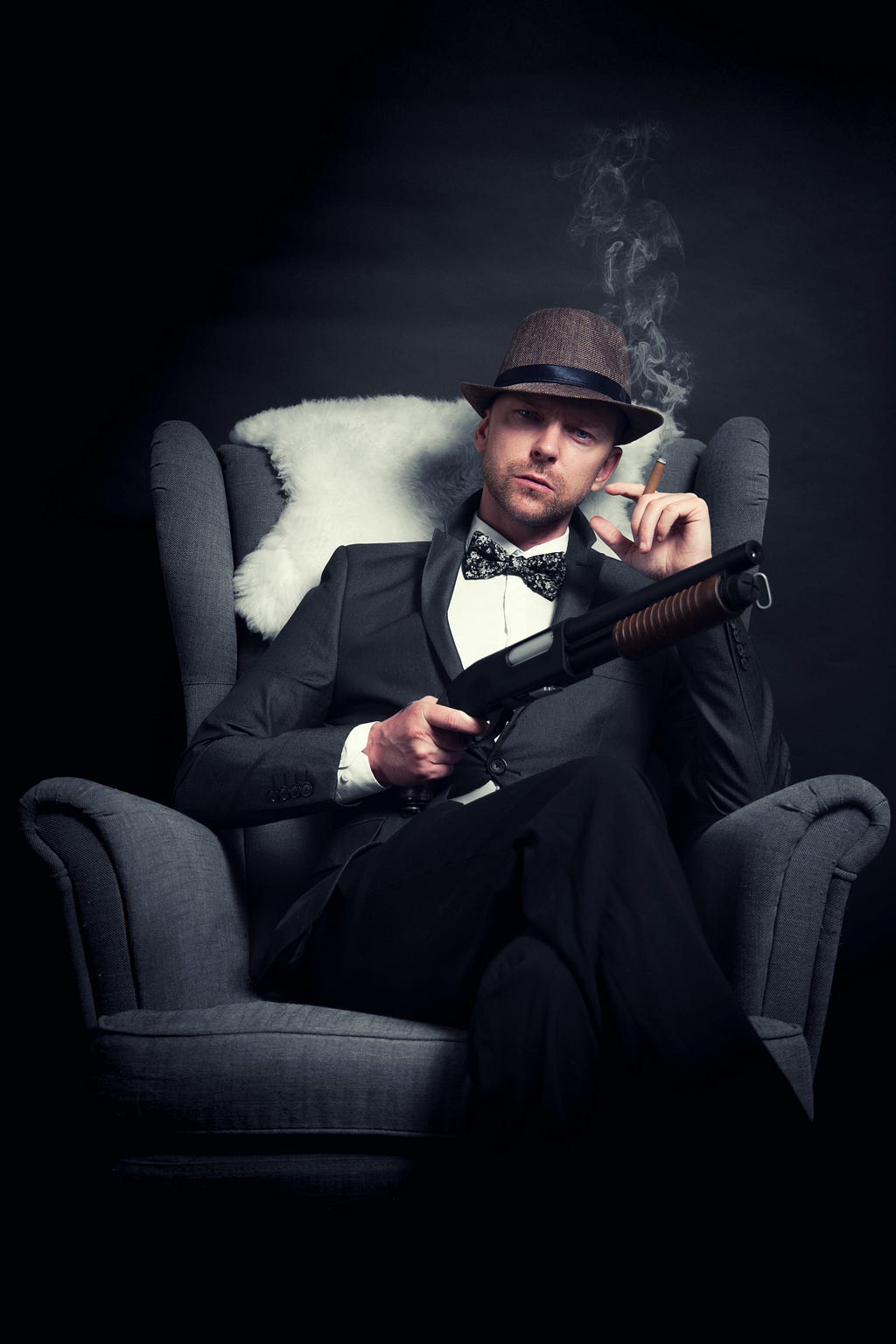 man with mobster appearance wearing old-time hat, bowtie and suit leaning in an armchair, holding a smoky cigarette in one hand and a type of shotgun in the other