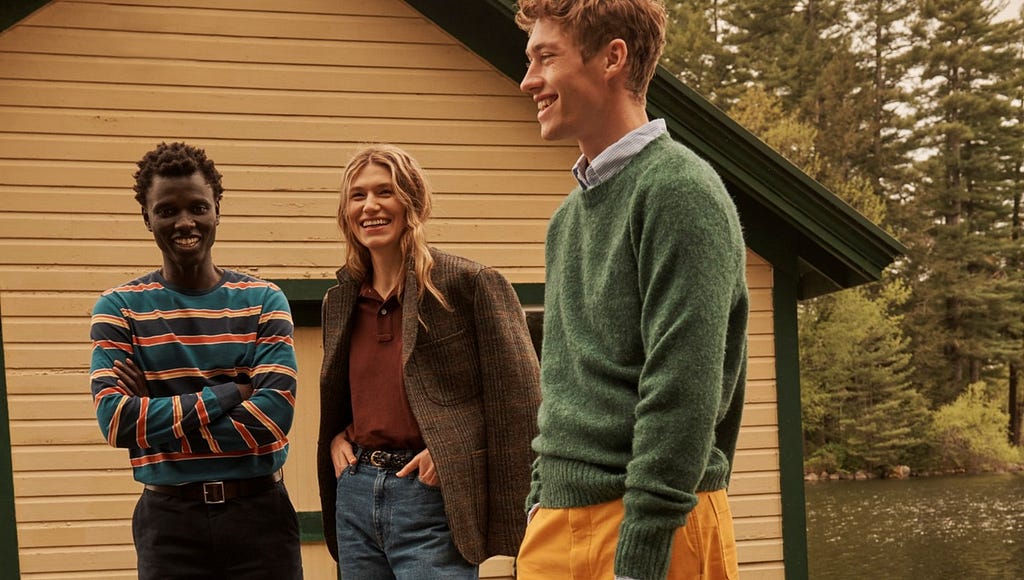 two men and one woman wearing autumnal clothing with bright colours — browns, greens, reds and oranges, standing next to a house near a lake — it feels like autumn/winter and they are smiling and chatting.