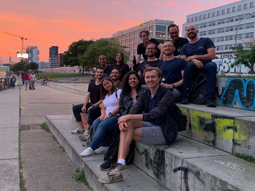 A photograph of 9 men and 3 women seated on concrete stairs in front a Berlin sunset