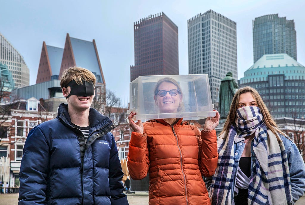 Jongen met een mondkapje over zijn ogen, vrouw met een doorzichtige doos op haar hoofd en meisje met een sjaal voor haar mond