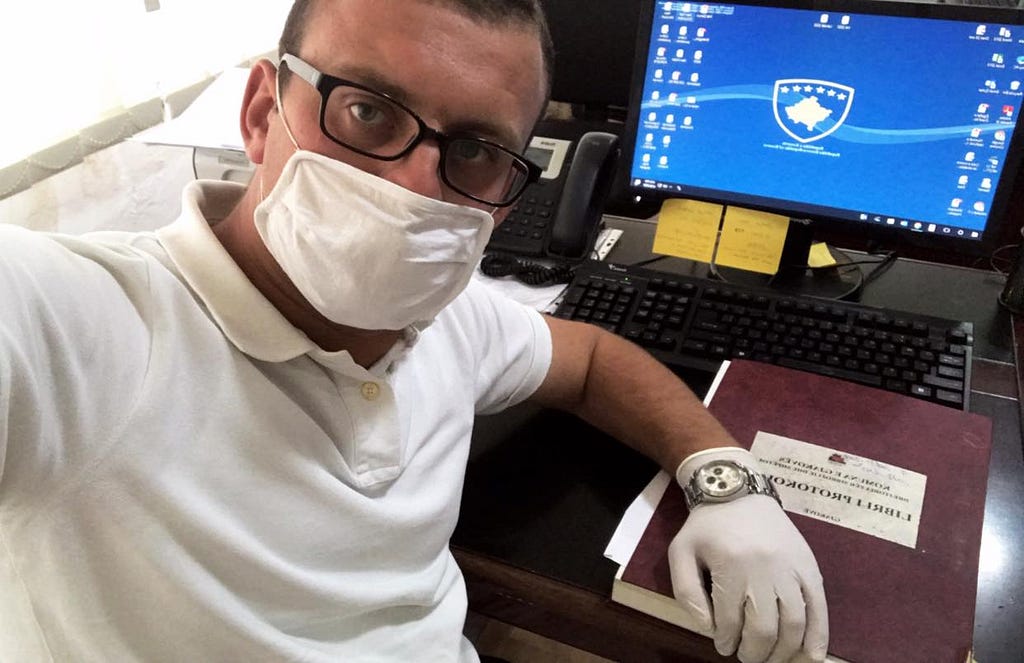 Besfort, wearing a white shirt, white facemask, white gloves, and glasses, looks at the camera, which he is holding