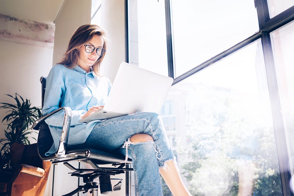 Young woman in glasses working on a laptop by a large window, wearing a blue shirt and ripped jeans, with natural light streaming in.