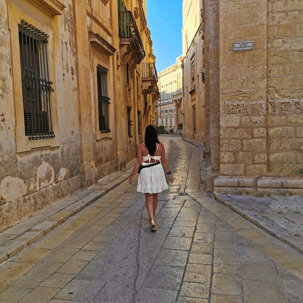 Victoria walking through the stone cobblestone streets of the Roman city Mdina (Medina), Malta during a warm day in August 2020. With dark brown hair and a white dress, admiring the beauty of the sandy stones that make up the narrow streets and tall stone buildings alike.