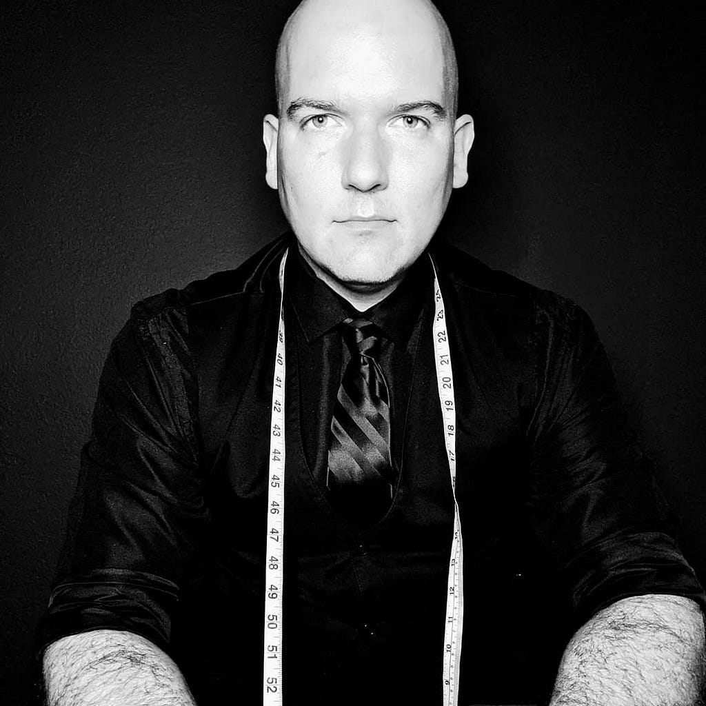 A white, bald man wearing a black shirt and tie stares intently at the camera with a measuring tape draped around his neck.