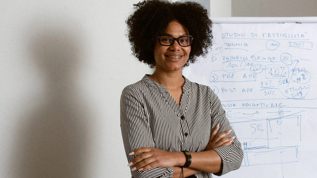 Black woman wearing glasses with curly afro standing with her arms crossed in front of a college chart.