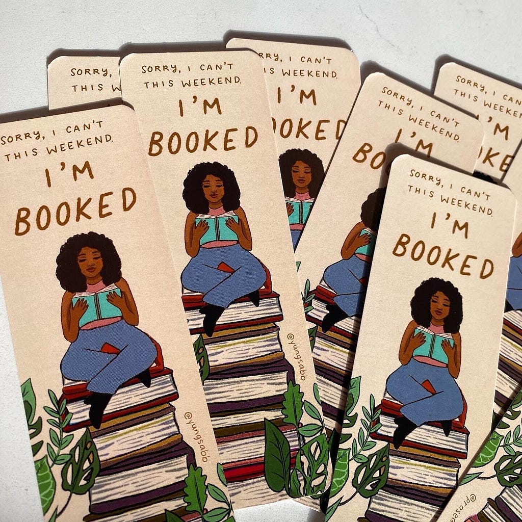 A bunch of bookmarks that say “Sorry I Can’t This Weekend, I’m Booked” on them. The bookmarks include a young woman sitting atop a stack of book of books reading.