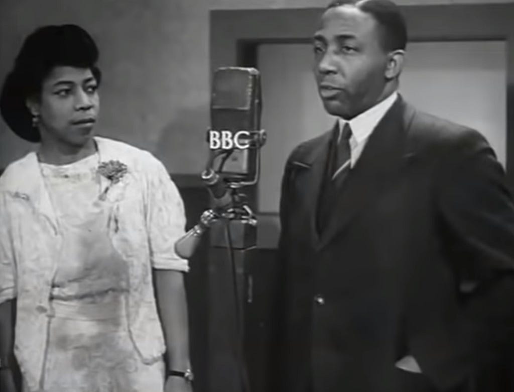 Black-and-white photograph of Una Marson (left) and Learie Constantine (right). Constantine is speaking into a microphone with the white letters BBC affixed on the front.