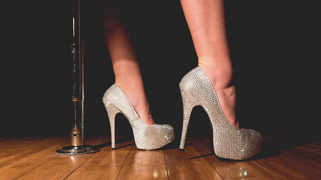 The feet of a woman wearing rhinestone studded white platform high-heels standing next to a stripper pole on a polished wooden plank stage.