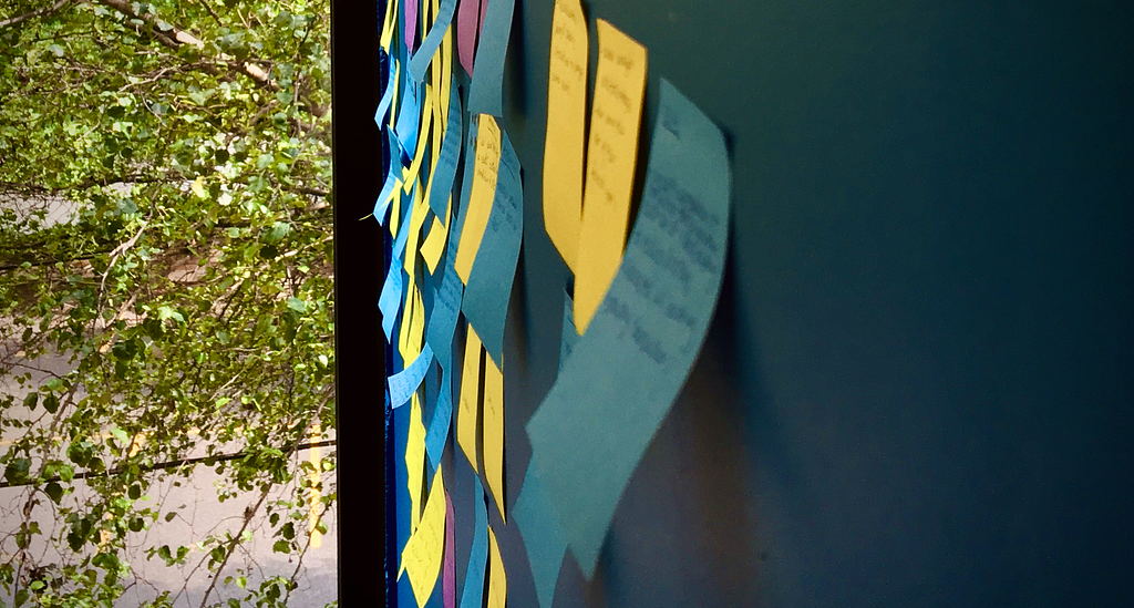 Post-It notes are organized in groups on a wall. A Spring green tree is visible through a window in the office.