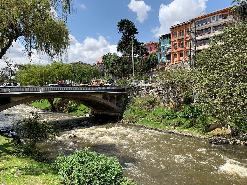 Tomebamba River of Cuenca. Pictured is a river rushing below a bridge; orange, blue and white buildings line the sidewalk.