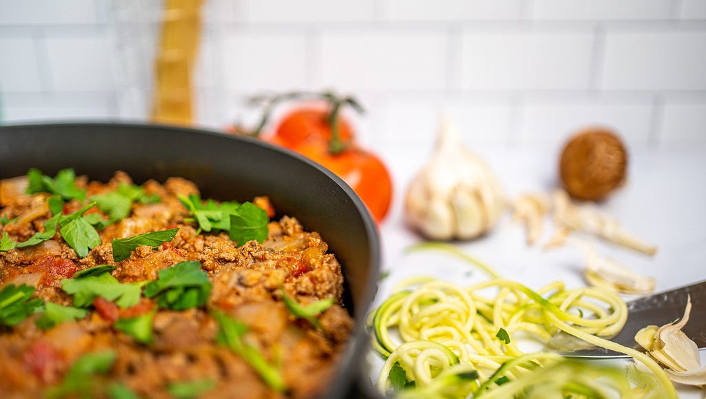 A large pan of cooked ground beef surrounded by garlic, zucchini, tomatoes, and parsley.