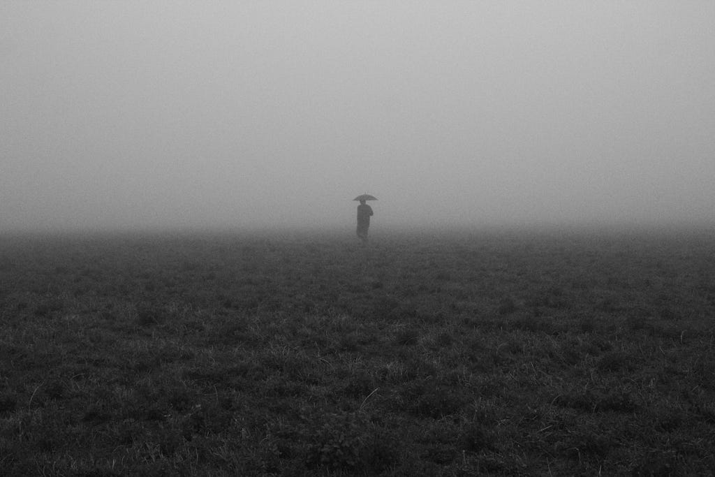 A black & white photo with a person standing on empty land, holding umbrellas, vanishing into the mist.