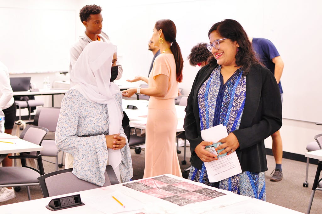 Photo of a teenage girl wearing a hijab talking to Mayor Sumbul Siddiqui about the t-shirt design sitting on the table in front of them. In the background, another student speaks with Cynthia Breazeal, and other students work on their own projects.