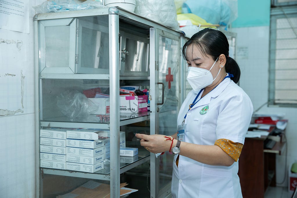 A health care worker stands beside shelves holding medicines and medical equipment.