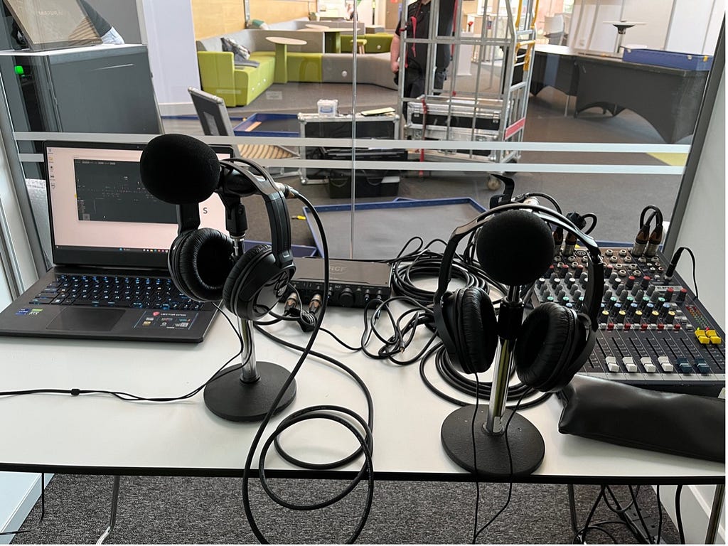 Photo of radio equipment on a table inside a glass-fronted booth. Includes a Windows laptop, two mic stands with mics and headphones on them, an audio interface and a small mixing desk. Behind the glass are scenes from a conference exhibition space being set up.