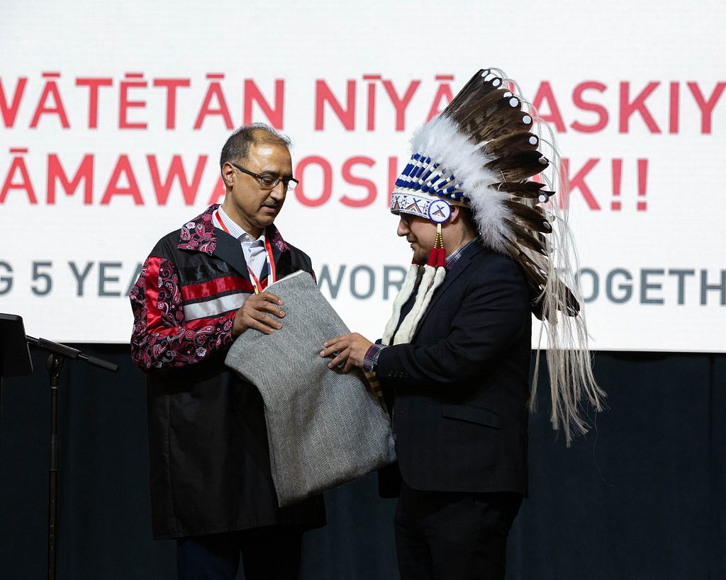 Chief Morin wears an eagle feather headdress and Sohi wears a red and black ribbon shirt as they unfold a ceremonial blanket.