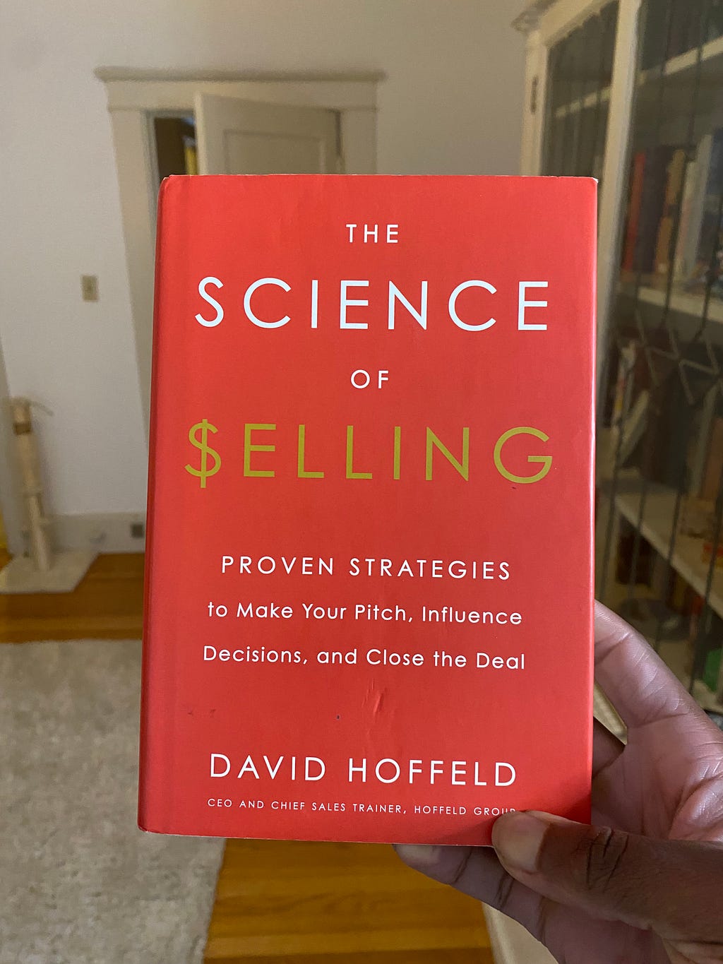 Hand holding up book in living room. Science of Selling by David Hoffeld