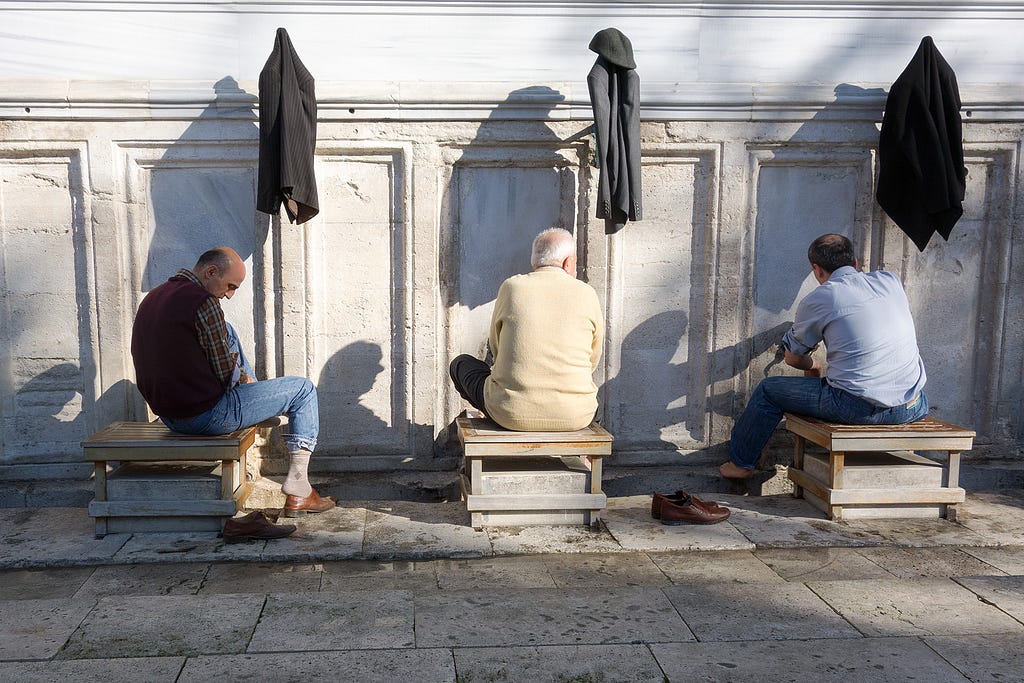 three men sitting on stools washing their feet at separate spigots