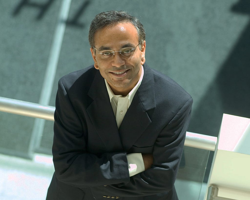 South Asian man in suit and glasses sits with crossed arms, looking up at the camera and smiling