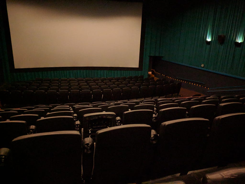 Interior of a nice movie theater from the back. Seats are brown leather; the walls are draped with teal green curtains. Sconces on the walls project light upwards. The screen is lit from above and blank (for now). The photo is REAL, taken by the article’s author.