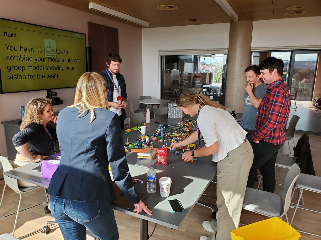 A group of people collaborating on a team project using Lego bricks. Some are standing and discussing, while others are seated around a table assembling models. The scene shows an interactive workspace with instructions projected on the wall.