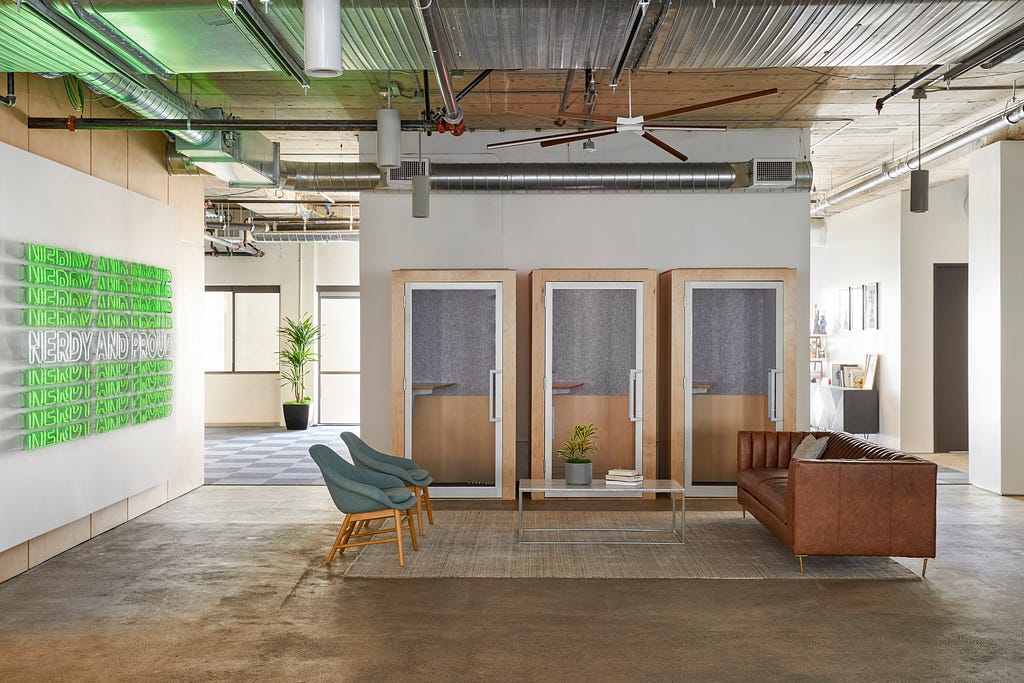 A zoomed out view of one of the spaces in the NerdWallet office. The “Nerdy and Proud” sign is on the left. In the middle, there are three phone booths, two chairs, and a couch.