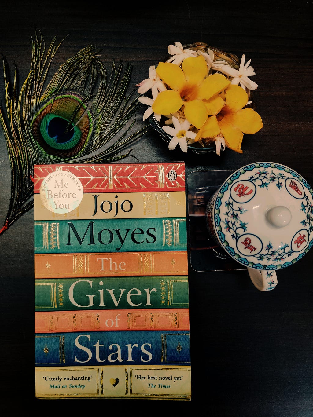 On my study table, an almost-square-ish arrangement of a book (“The Giver of Starts” by Jojo Moyes) along the left margin, a peacock feather tilted on the top left corner, a fresh white and yellow flower arrangement on the top right corner and a cup of tea along the right margin.