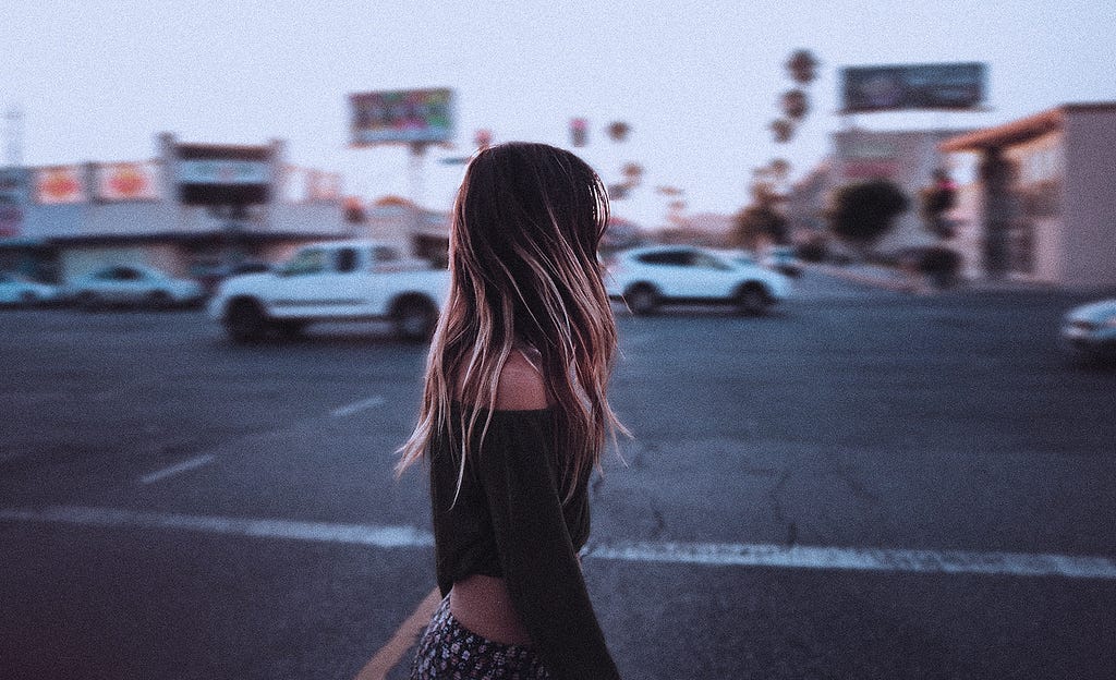 A girl is walking in a city covering her face with her blond hair, few cars are passing on the road