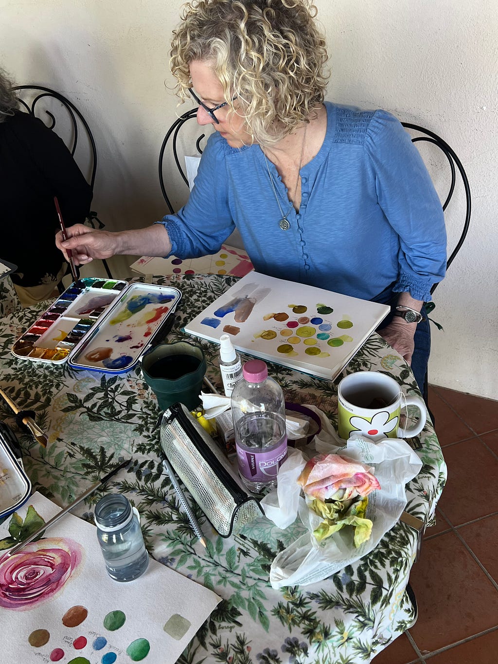 Watercolor artist Roxanne Steed sits at a table spread with palettes, paper, color wheels, and water.