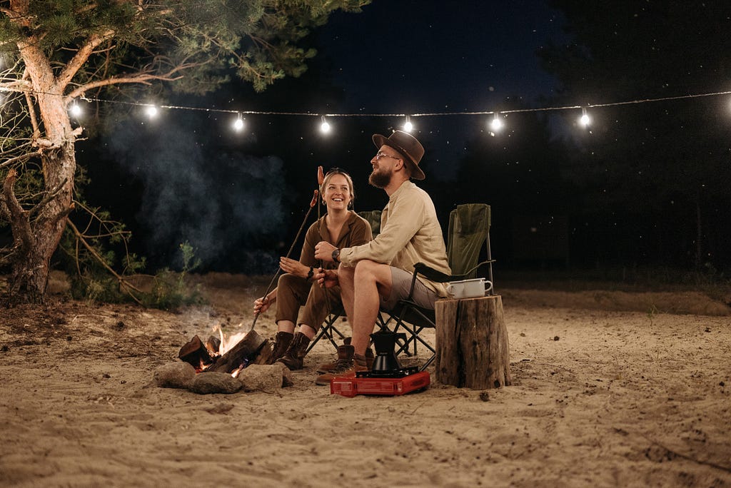 Couple sitting on chairs while grilling on campfire.