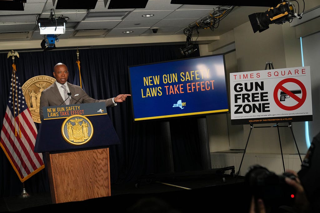 Mayor Adams stands at a podium, pointing to two signs, one reading “New Gun Safety Laws Take Effect” and the other reading “Times Square Gun Free Zone”