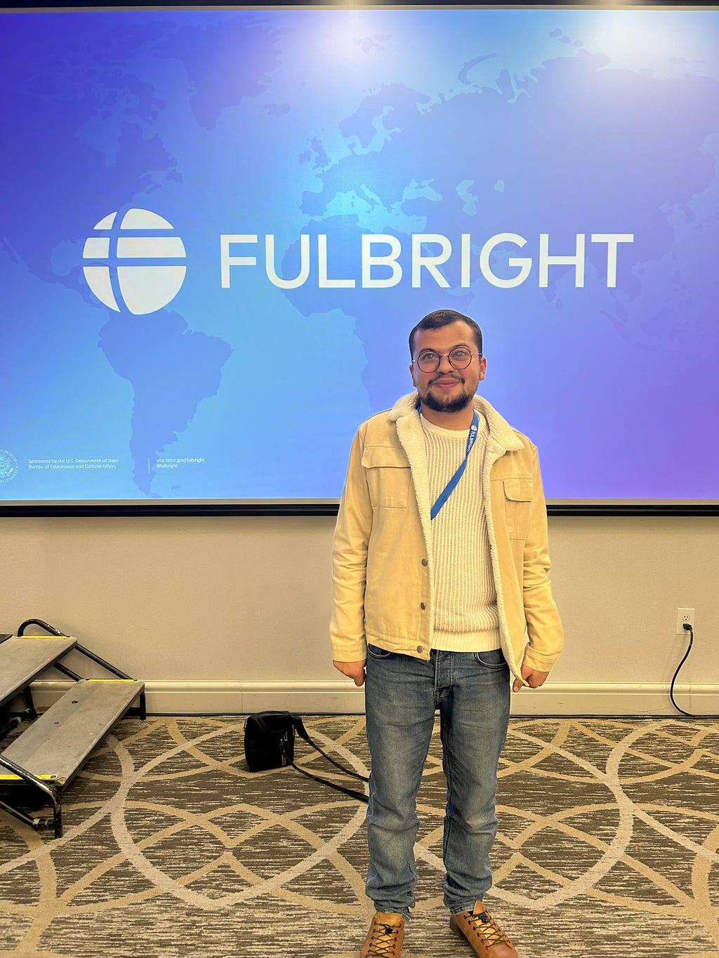 Mu’nis in a cream-colored sweater and jacket standing before a blue screen featuring the Fulbright logo.