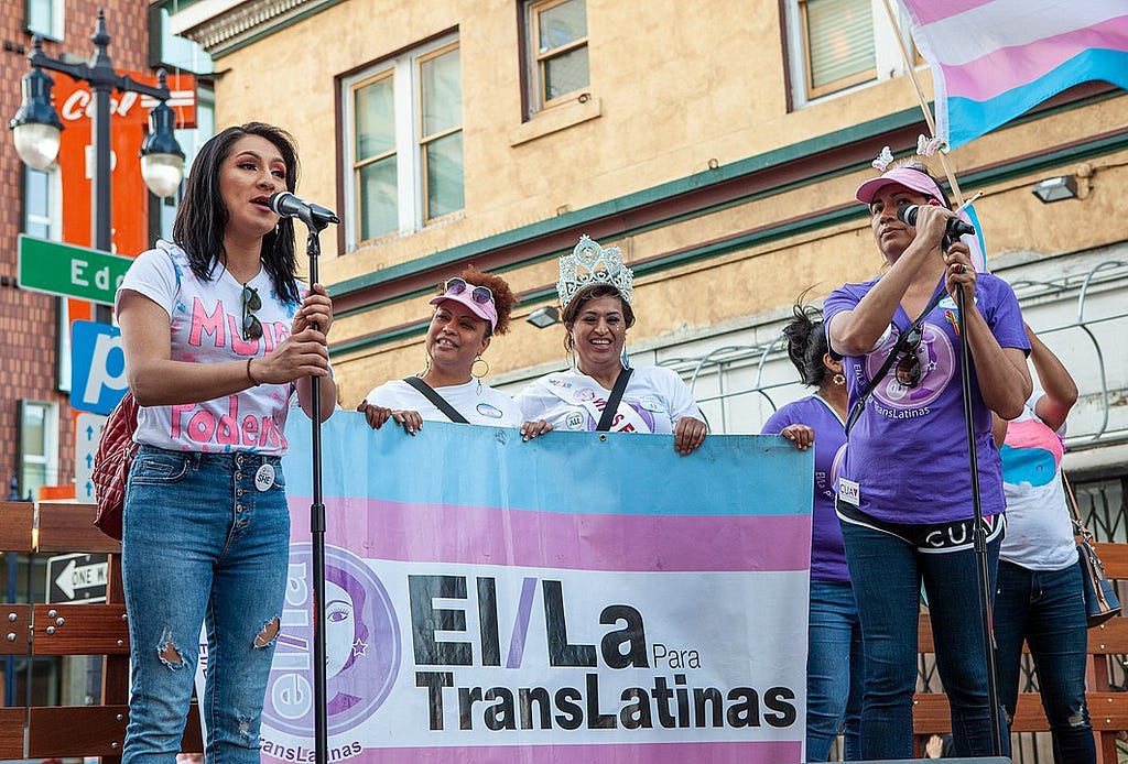 A group of trans Latinas stand on a stage. One speaks into a mic, one holds a trans flag, others hold a banner.