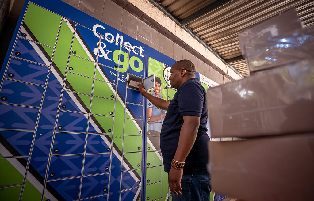 A man reaches into a secure package locker, also known as a “smart locker,” to remove a box. The locker is brightly colored and blue and green with the company’s logo — Collect & Go™.