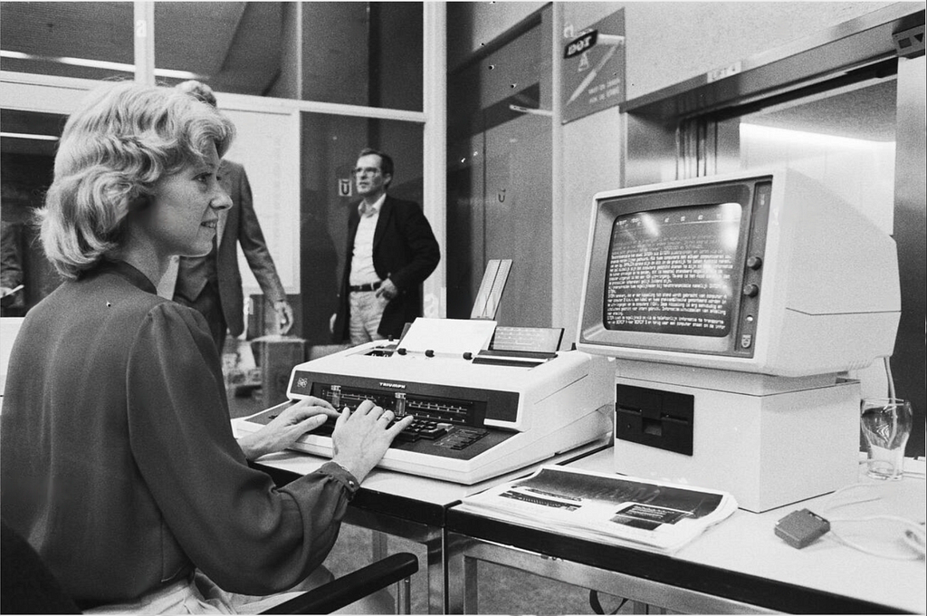 A black-and-white photograph of a woman seated at a desk working on a vintage computer system. The setup includes a large CRT monitor displaying text, a typewriter-style keyboard with paper being fed through it, and additional documents or equipment on the desk. In the background, two men are standing and talking near a glass partition. The scene appears to be from an office or exhibition setting